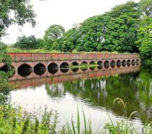 A scenic view of the historic brick bridge with multiple arches spanning across the tranquil river at Carr Mill Dam - St Helens. The bridge and lush green trees are beautifully reflected in the calm water, creating a serene and picturesque landscape.