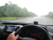 A person is driving a car on a highway. The dashboard and steering wheel are visible, with a GPS navigation system displaying directions. The road is slightly wet, suggesting recent rain, and a few cars are ahead. Lush green trees line the road on the left side.