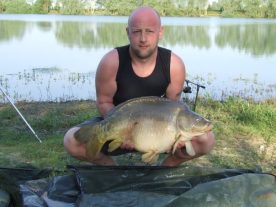 A person is kneeling on a mat beside a body of water, holding a large fish with both hands. The person is wearing a black sleeveless shirt and shorts, with fishing equipment visible in the background. Trees and a calm lake are also in the background.