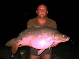 A person is standing outdoors at night holding a large fish. The person has a serious expression and is wearing a brown shirt and shorts. The background is dark, making the fish, which is a light color, stand out prominently.