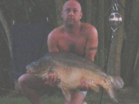 A person sits shirtless on a chair outdoors, holding a large fish in their hands. There is a fishing rod with a bell attached standing next to them, and trees are visible in the background. The scene appears to be at dusk.