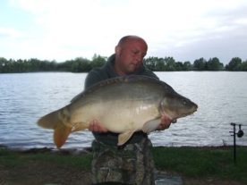 A person wearing a green hoodie and camouflage pants is holding a large fish in front of a lake. The background shows trees and a cloudy sky. Fishing gear is visible on the ground and near the water's edge.