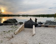 A peaceful lakeside campsite at sunset with three tents, fishing gear, and two people standing and talking. The sky displays beautiful evening colors, and the still water of Pegasus Lakes in France reflects the scene, creating a tranquil atmosphere.