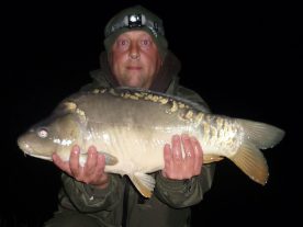 A person wearing dark outdoor clothing and a headlamp holds a large fish, possibly a carp, with both hands. The background is dark, suggesting the photo was taken at night, making it an ideal shot for a Beginners Guide to Night Fishing for Carp.