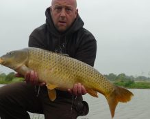 A person in a dark hoodie kneels by a body of water at Cool Acres, holding a large golden-brown fish with a slightly surprised expression. The background features overcast skies and distant trees, suggesting a peaceful outdoor fishing scene in the UK.