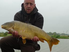 A person with a shaved head and wearing a dark hoodie holds a large, golden-colored fish while kneeling near a peaceful lakeside. The sky is overcast, and greenery lines the shore in the background.