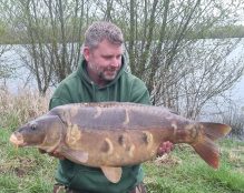 A man in a green hoodie, crouching on a grassy bank beside Pegasus Lakes - France, holds a large, brown carp fish. The background has leafless, slender trees and calm water, suggesting early spring. The man looks pleased as he gazes at the fish.