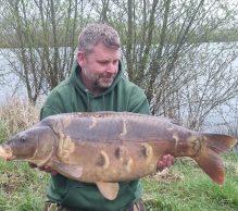 A man in a green hoodie, crouching on a grassy bank beside Pegasus Lakes - France, holds a large, brown carp fish. The background has leafless, slender trees and calm water, suggesting early spring. The man looks pleased as he gazes at the fish.