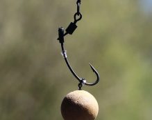 A close-up of a fishing hook hanging vertically, with a round bait attached at the bottom. The background is blurred, showcasing a mix of green and brown hues, indicating an outdoor environment near water—perfect for making a 360 carp rig.