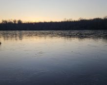 A serene, frozen lake at dusk is bordered by a forest of leafless trees. The sun is setting on the horizon, casting a soft, golden glow over the tranquil scene. The glassy surface of the ice reflects the fading light, highlighting intricate patterns—a hidden gem for carp anglers like Etang Rapide.