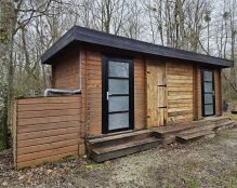 A small wooden cabin with two doors and two large windows. The building has a flat, black-topped roof and is situated in a forested area with bare trees near Etang Rapide, a hidden gem for carp anglers in France. A wooden platform with steps leads to the front entrances, and a fenced area is attached on the left side.