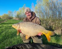 A person, dressed in a brown hoodie and beanie, is kneeling on the grass at Pegasus Lakes - France and holding a large carp fish with both hands. Trees and blue sky form the tranquil backdrop. The scene is outdoors, with bright sunlight casting shadows.