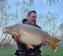 A man in camouflage clothing smiles while holding a large carp fish with both hands at Pegasus Lakes - France. He is outdoors with trees and a green field visible in the background. It appears to be a pleasant, clear day.