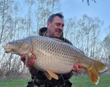 A man in camouflage clothing smiles while holding a large carp fish with both hands at Pegasus Lakes - France. He is outdoors with trees and a green field visible in the background. It appears to be a pleasant, clear day.