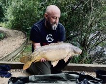 A bearded man wearing a black t-shirt and dark green pants is kneeling beside a small pond surrounded by lush greenery. He is holding a large carp fish with both hands, showing it off proudly. In the background, there is a dirt pathway and dense foliage.