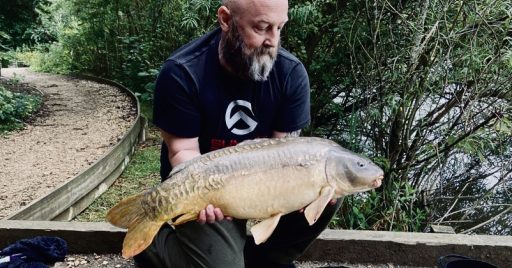 A bearded man wearing a black t-shirt and dark green pants is kneeling beside a small pond surrounded by lush greenery. He is holding a large carp fish with both hands, showing it off proudly. In the background, there is a dirt pathway and dense foliage.