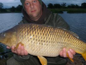 A person in green winter clothing and a hat poses by a lake, holding a large, golden-colored fish with shiny scales. The background includes trees and a cloudy sky.