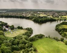 Aerial view of Carr Mill Dam - St Helens, surrounded by lush greenery and trees. A small gathering with tents and vehicles is visible on the left shore, near a railway track. Fields and residential areas stretch out in the background under an overcast sky.