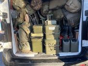 The back of a white van filled with various outdoor gear and military-style equipment, including green bags, rolled-up tents or tarps, two green metal containers, black boots, and two green fuel cans. The van is parked on a paved surface.