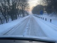 View from inside a car driving on a snow-covered road lined with bare trees on both sides. The sky is a mix of soft blue and pink hues, indicating dawn or dusk. The road appears slippery, and a blue road sign is visible in the distance on the right side.