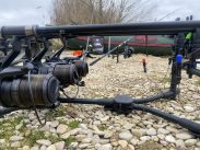 Close-up of fishing rods set up on a rocky lakeshore, with a green inflatable fishing boat and trees in the background. The rods are mounted on a black holder, and lines are extended toward the water, suggesting a fishing trip in progress.