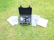 A black hard-shell case with foam padding is open on a grassy surface. Inside the case are various tools and components neatly organized. Surrounding the case are four transparent plastic bags, each containing additional parts, and a white piece of paper.