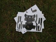 Stickers resembling parts of a suit of armor with a camouflage pattern are arranged on several white sheets of paper placed on grass.