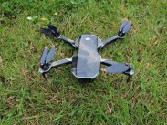 A small, camo-colored drone with four propellers sits on a patch of green grass. The drone's compact design features black and gray colors, with orange accents on the propellers. The grass around the drone is lush and evenly cut.