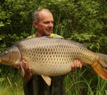 A man wearing dark waterproof overalls is standing outdoors, holding a large carp fish. The background is lush and green with tall grass and trees, suggesting a natural, freshwater setting. The man appears to be proud of his catch at Etang Rapide, a hidden gem for carp anglers in France.