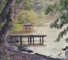 A serene lakeside scene with two wooden piers extending into the calm water at Carr Mill Dam - St Helens. The shoreline is dotted with lush green trees, creating a peaceful, natural atmosphere. The overcast sky adds a muted, tranquil vibe to the setting.