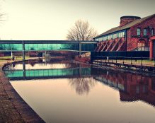 A serene canal reflects a historic industrial brick building with arched windows and a modern glass walkway. Leafless trees stand on the path alongside, contrasting old and new architectural elements in a tranquil, overcast setting at The Hotties - St Helens canal.