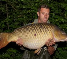 A person stands in front of dense green foliage, holding a very large fish with both hands. The fish has a golden-brown scale pattern and a long, slightly tapered tail. The person is gazing at the fish while wearing an outdoor jacket at Etang Rapide, a hidden gem for carp anglers in France.