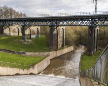 A river flows under a steel railway bridge where a few birds are perched. The river narrows between concrete walls below the bridge. On both sides of the river are grassy areas, with some graffiti visible on the bridge supports, reminiscent of scenes around Carr Mill Dam - St Helens. Trees are visible in the background.