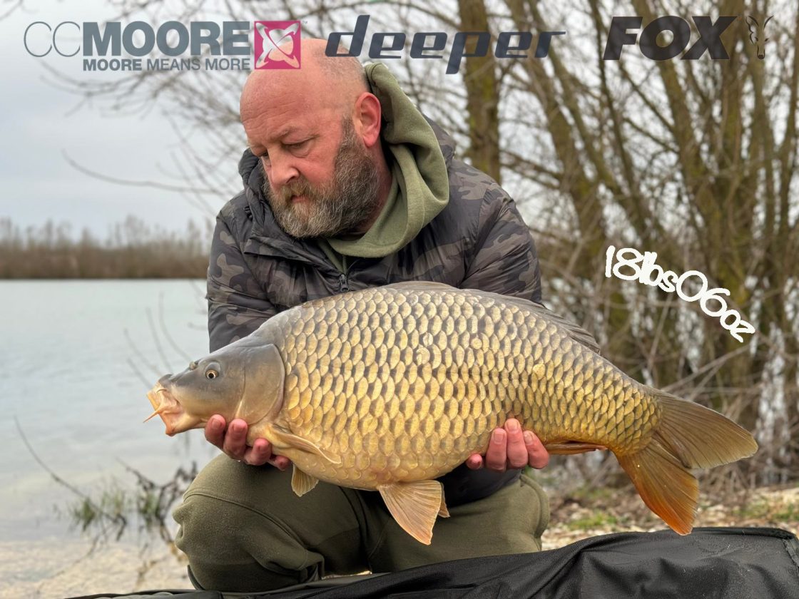 A person with a beard, dressed in camouflage gear, is holding a large, golden fish near a lake. The fish's weight, 