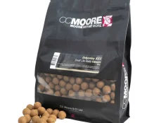 A black bag of CC Moore Odyssey XXX Shelf Life Baits 15mm is depicted. The bag, essential for those looking to bait up in pressured waters, has a transparent section revealing round brown bait balls, some of which are scattered on the white surface next to the bag.