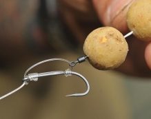 A close-up of a person's hand holding a fishing rig, featuring a sharp fish hook and two round bait balls threaded onto the line. The intricacy of tying a D rig and the texture of the bait are clearly visible, showcasing when to use it for optimal results.