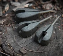 Close-up of three dark gray fishing weights lying on a weathered tree stump surrounded by dry leaves and twigs. The solid weights are elongated with a narrow slit running along their length and a pointed end, perfect for rigging a simple PVA bag setup.