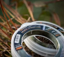 Close-up of a bunch of packaged double-tapered fishing lines by an outdoor setting with dry vegetation in the background. The transparent packages, revealing braided fishing lines inside, partially show the brand name 