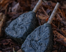 Close-up image of two teardrop-shaped fishing sinkers, similar to carp leads, on a ground covered with brown mulch. The sinkers, marked with 