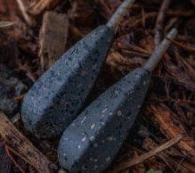 Two black, stone-like arrowheads with speckled patterns rest side by side on a bed of wood chips and natural debris, reminiscent of carp leads. The arrowheads have a tapered shape with a short stem extending from their wider ends. A small green leaf is visible at the bottom right corner.