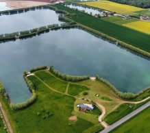 Aerial view of Pegasus Lakes - France showcases a rural landscape with several large, rectangular ponds surrounded by green fields and trees. A small building and pathway are visible near the bottom of the image, adjacent to the ponds, while a road runs along the right edge.