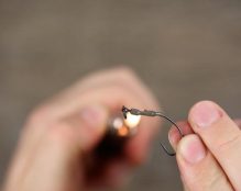 A close-up of hands holding a fishing hook with a small metal piece attached, demonstrating one of the 50 commonly used carp rigs. One hand is holding the hook while the other uses a lit lighter to heat something on it. The background is blurred.