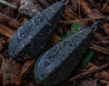 Two teardrop-shaped carp leads lie on a bed of mulch and twigs. The weights, marked 